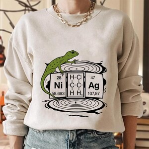 May include: Beige sweatshirt with a green lizard design above a graphic resembling a periodic table, with elements Ni and Ag. The graphic also includes the text "H-C-C-H-H".
