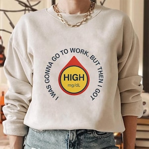 May include: Beige sweatshirt featuring the text "I WAS GONNA GO TO WORK, BUT THEN I GOT HIGH mg/dL" around a red and yellow graphic. The graphic is a blood drop shape with the word "HIGH" inside.