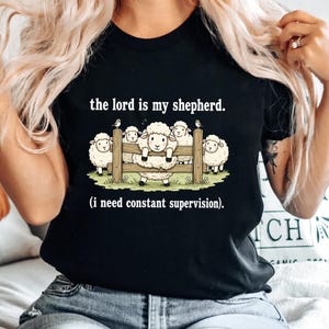 May include: Black t-shirt featuring a cartoon illustration of sheep behind a wooden fence. The text "the lord is my shepherd. (i need constant supervision)." is printed above and below the sheep graphic. The shirt is a casual wear item.