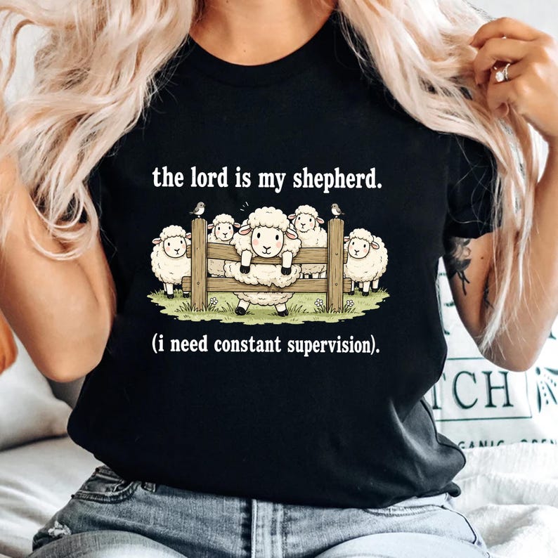 May include: Black t-shirt featuring a cartoon illustration of sheep behind a wooden fence. The text "the lord is my shepherd. (i need constant supervision)." is printed above and below the sheep graphic. The shirt is a casual wear item.