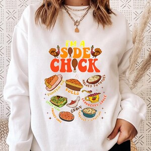 May include: White sweatshirt with a colorful graphic that reads "I'M A SIDE CHICK" with images of Thanksgiving side dishes. The design includes pumpkin pie, mashed potatoes, rolls, and cranberry sauce.