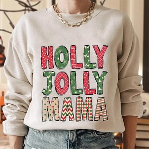 May include: A beige sweatshirt with the words "HOLLY JOLLY MAMA" in a festive, Christmas-themed design. The letters are in red and green with snowflake, reindeer, and patterned accents. A gold chain necklace is visible.