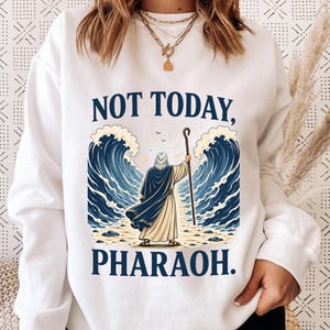 Könnte beinhalten: Weißes Sweatshirt mit dem Text "NOT TODAY, PHARAOH." Die Grafik zeigt eine Person mit einem Stab zwischen zwei großen blauen Wellen. Die Person trägt einen blauen Umhang und hält einen Stab.