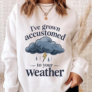May include: A white long-sleeve sweatshirt featuring a dark blue graphic of a storm cloud with a yellow lightning bolt and raindrops. The text reads I've grown accustomed to your weather in dark blue lettering across the front.