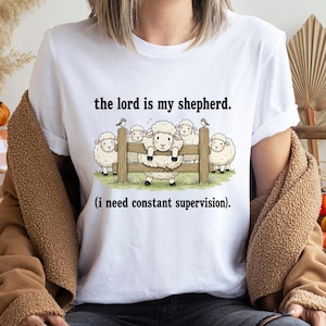 May include: White t-shirt with the text "the lord is my shepherd" above a cartoon illustration of sheep behind a wooden fence. The text "(i need constant supervision)" is below the illustration.