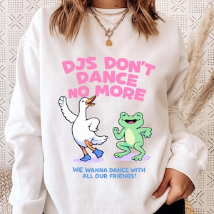 May include: White sweatshirt featuring the pink text "DJs Don't Dance No More". The design includes cartoon illustrations of a dancing duck and frog. The phrase "We Wanna Dance With All Our Friends!" is printed below the illustrations.