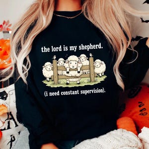 May include: Black sweatshirt with the text "the lord is my shepherd." and a graphic of sheep behind a wooden fence. The phrase "(i need constant supervision)" is below the graphic.