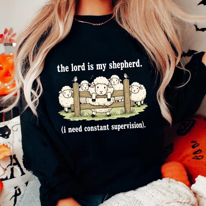 May include: Black sweatshirt with the text "the lord is my shepherd." and a graphic of sheep behind a wooden fence. The phrase "(i need constant supervision)" is below the graphic.