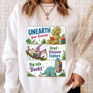 Op de afbeelding: Witte sweatshirt met drie rechthoekige afbeeldingen. Elke afbeelding bevat een dinosaurusillustratie en tekst. De bovenste afbeelding luidt "Unearth Your Summer". De middelste afbeelding zegt "Read Discover Explore". De onderste afbeelding zegt "Dig into Books."