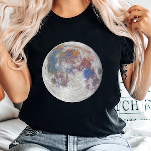 May include: A black short-sleeved t-shirt featuring a large, circular graphic of the moon on the chest. The moon illustration displays a vibrant, multi-colored surface with distinct patches of blue, orange, and purple hues against a gray base.