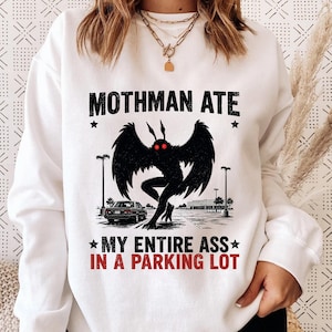 May include: A white long-sleeve sweatshirt featuring a black graphic of the Mothman with red eyes standing in a parking lot. The text reads "MOTHMAN ATE" in black and "MY ENTIRE ASS IN A PARKING LOT" in red, accented by two black stars.