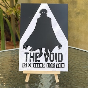 May include: A rectangular print featuring a black silhouette of a figure in a cape with glowing white eyes, set against a white triangular background. The text THE VOID IS CALLING FOR YOU is printed in bold black capital letters at the bottom.