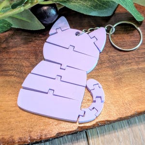 Op de afbeelding: Een lichtpaarse sleutelhanger in de vorm van een kat met een gesegmenteerd, gearticuleerd lichaamsontwerp. De katfiguur is opgebouwd uit verschillende horizontale delen die verbonden zijn door scharnieren, inclusief een gebogen staart.