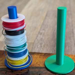 Può includere: Una pila di cinque bobine di plastica avvolte con filo elettrico rosso, nero, bianco, verde, blu e giallo, tenute su un supporto verticale blu, accanto a un supporto verticale verde vuoto con base circolare.