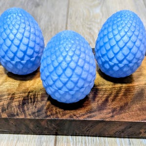 May include: Three light blue, egg-shaped objects with a textured, scale-like pattern, resting on a dark wooden surface. The eggs are evenly spaced, creating a symmetrical composition. The wood grain adds a natural contrast to the smooth eggs.