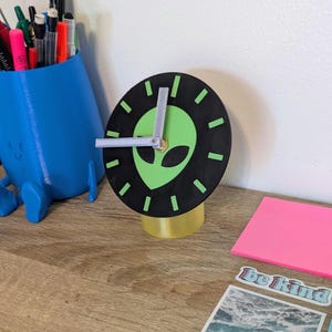 May include: A black and green alien-themed desk clock. The clock face features a green alien head on a black background with green hour markers. The clock hands are silver, and the base is yellow. A blue pen holder and sticky notes are in the background.