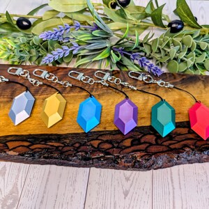 Peut inclure: Six porte-clés en forme de pierres précieuses, argent, or, bleu, violet, vert d'eau et rouge. Chaque porte-clés a un fermoir argenté et un cordon noir. Ils sont présentés sur une surface en bois, avec des plantes en arrière-plan.