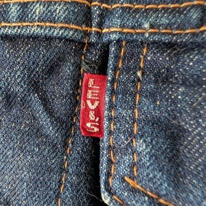 May include: Close-up of dark blue denim fabric with orange stitching. A small red tag with white text is attached to the denim. The stitching is visible along the seams and edges of the fabric.