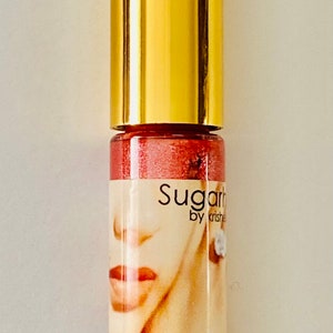 Puede incluir: Un tubo dorado de brillo de labios Sugar by Krishe con un acabado rojo brillante. El tubo tiene una etiqueta con el rostro de una mujer y el texto "Sugar by Krishe" y "Lip".