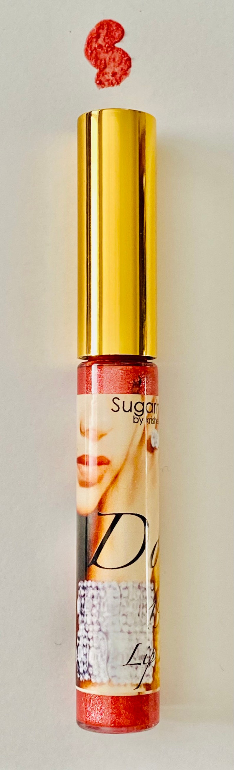 Puede incluir: Un tubo dorado de brillo de labios Sugar by Krishe con un acabado rojo brillante. El tubo tiene una etiqueta con el rostro de una mujer y el texto "Sugar by Krishe" y "Lip".