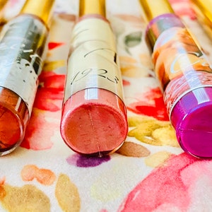 Puede incluir: Seis tubos transparentes de brillo de labios con tapas doradas. Los tubos tienen etiquetas de diferentes colores y los brillos de labios son visibles a trav&eacute;s de los tubos transparentes. Los colores de brillo de labios son marr&oacute;n, rosa, rosa claro, morado y naranja.