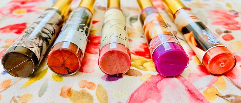 Puede incluir: Seis tubos transparentes de brillo de labios con tapas doradas. Los tubos tienen etiquetas de diferentes colores y los brillos de labios son visibles a trav&eacute;s de los tubos transparentes. Los colores de brillo de labios son marr&oacute;n, rosa, rosa claro, morado y naranja.