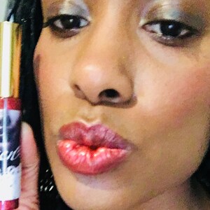 Puede incluir: Primer plano de los labios de una persona que lleva un brillo de labios rojo transparente. El brillo es brillante y tiene un ligero brillo. La persona sonr&iacute;e y tiene un aspecto de maquillaje natural.