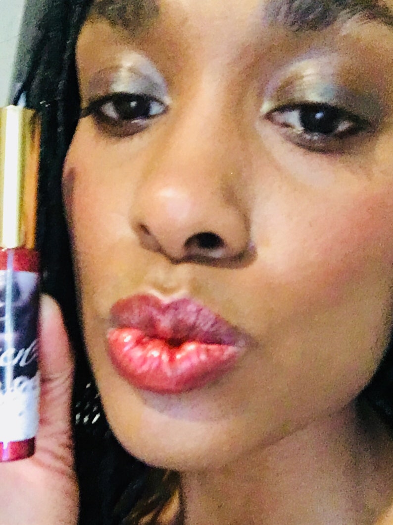 Puede incluir: Primer plano de los labios de una persona que lleva un brillo de labios rojo transparente. El brillo es brillante y tiene un ligero brillo. La persona sonr&iacute;e y tiene un aspecto de maquillaje natural.