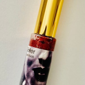 Puede incluir: Un tubo dorado con un brillo de labios rojo con purpurina en su interior. El tubo tiene una etiqueta con el texto "Sister" y "Essentials". El brillo de labios es de color rojo intenso con un acabado brillante.