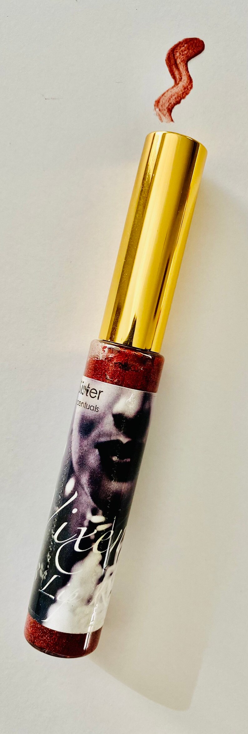 Puede incluir: Un tubo dorado con un brillo de labios rojo con purpurina en su interior. El tubo tiene una etiqueta con el texto "Sister" y "Essentials". El brillo de labios es de color rojo intenso con un acabado brillante.