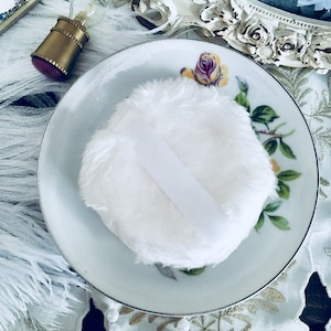 May include: A white fluffy powder puff with a white satin ribbon on a white porcelain saucer with a floral design. The saucer is on a white lace tablecloth with a pearl necklace.