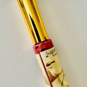 Puede incluir: Un tubo dorado de brillo de labios Sugarfina con un l&iacute;quido rojo brillante en su interior. El tubo tiene una etiqueta con el texto "Sugarfina by Kendra Scott" y el n&uacute;mero "1922".