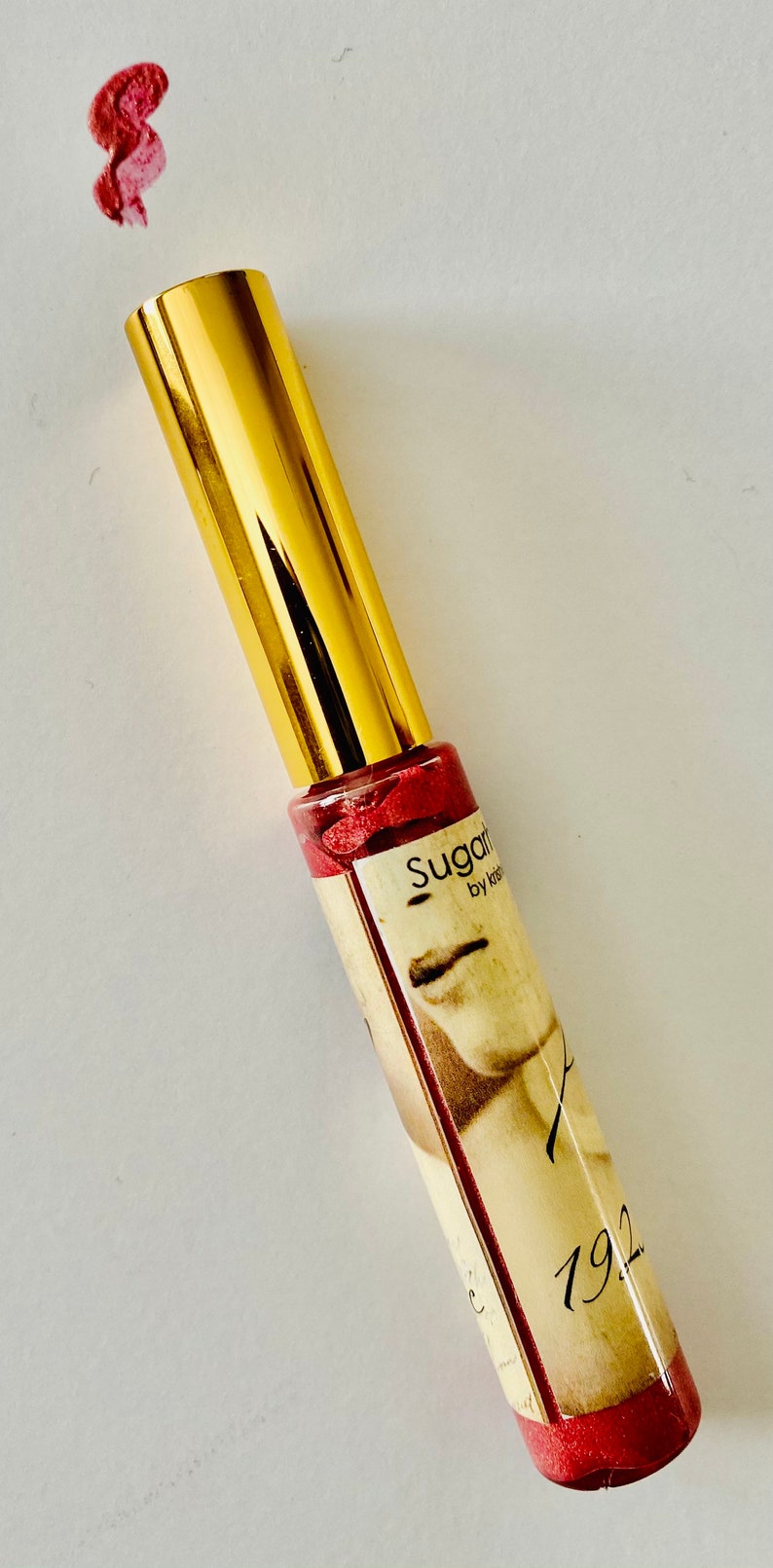 Puede incluir: Un tubo dorado de brillo de labios Sugarfina con un l&iacute;quido rojo brillante en su interior. El tubo tiene una etiqueta con el texto "Sugarfina by Kendra Scott" y el n&uacute;mero "1922".