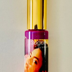 Puede incluir: Un tubo de brillo de labios dorado y morado con una etiqueta que dice "Sugarpill by Kristee" y una imagen de la cara de una mujer. El brillo de labios es de color morado brillante.
