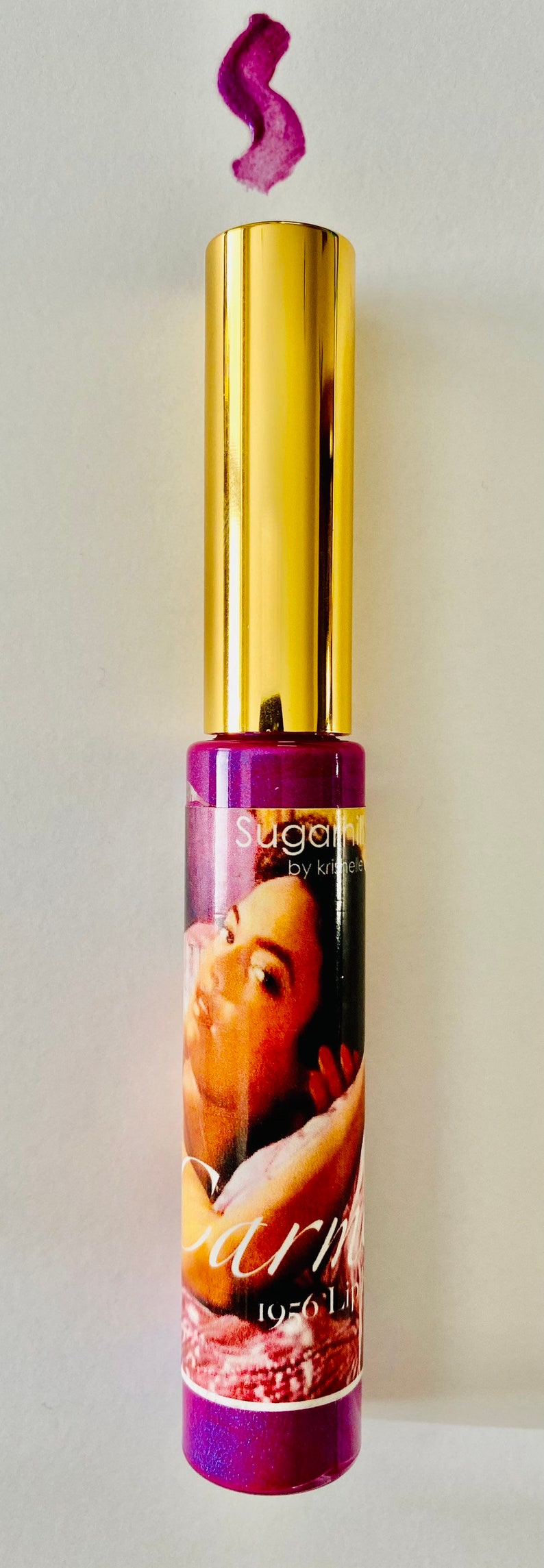 Puede incluir: Un tubo de brillo de labios dorado y morado con una etiqueta que dice "Sugarpill by Kristee" y una imagen de la cara de una mujer. El brillo de labios es de color morado brillante.