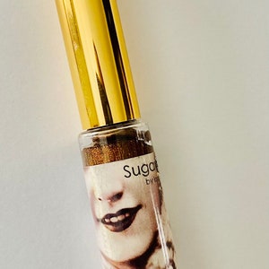 Puede incluir: Un tubo dorado con una etiqueta en blanco y negro que dice "Sugar by Kylie" y presenta el rostro de una mujer. El tubo est&aacute; sobre un fondo blanco.