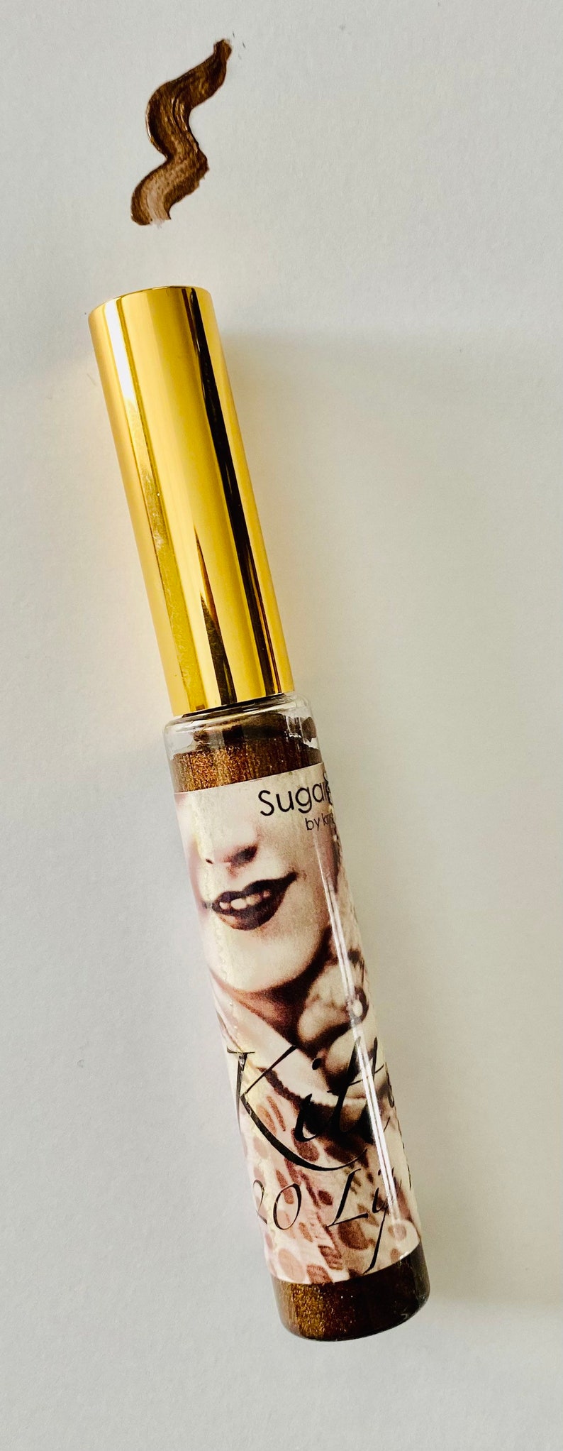 Puede incluir: Un tubo dorado con una etiqueta en blanco y negro que dice "Sugar by Kylie" y presenta el rostro de una mujer. El tubo est&aacute; sobre un fondo blanco.