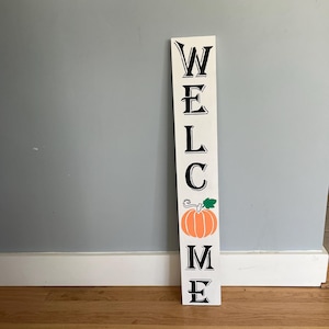 Op de afbeelding: Een wit houten welkomstbord met zwarte letters en een oranje pompoenillustratie. Het bord is verticaal en staat op een houten vloer, ideaal voor seizoensgebonden woondecoratie.