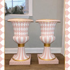 May include: Two matching decorative urns. Each urn features a pink and white harlequin pattern on the body, with gold accents at the base and rim. The base is square and pink with gold dots. The urns are set against a neutral background.