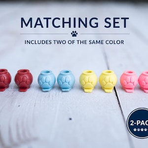 May include: A set of eight small, round, hollow silicone charms, each featuring a raised paw print design. The charms are arranged in pairs of red, light blue, yellow, and pink. Text reads MATCHING SET INCLUDES TWO OF THE SAME COLOR and 2-PACK.