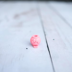 May include: A small, light pink silicone bead shaped like a rounded container with a central opening at the top. The exterior surface features a raised, embossed paw print design on the front, and the base has a flat, protruding tab extension.