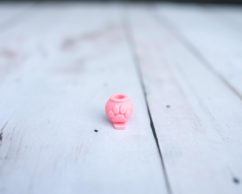 May include: A small, light pink silicone bead shaped like a rounded container with a central opening at the top. The exterior surface features a raised, embossed paw print design on the front, and the base has a flat, protruding tab extension.