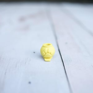 May include: A small, yellow, spherical bead featuring a raised paw print design on its front surface. The bead has a circular opening at the top and a small, flat, protruding base at the bottom, designed for threading onto a cord or string.