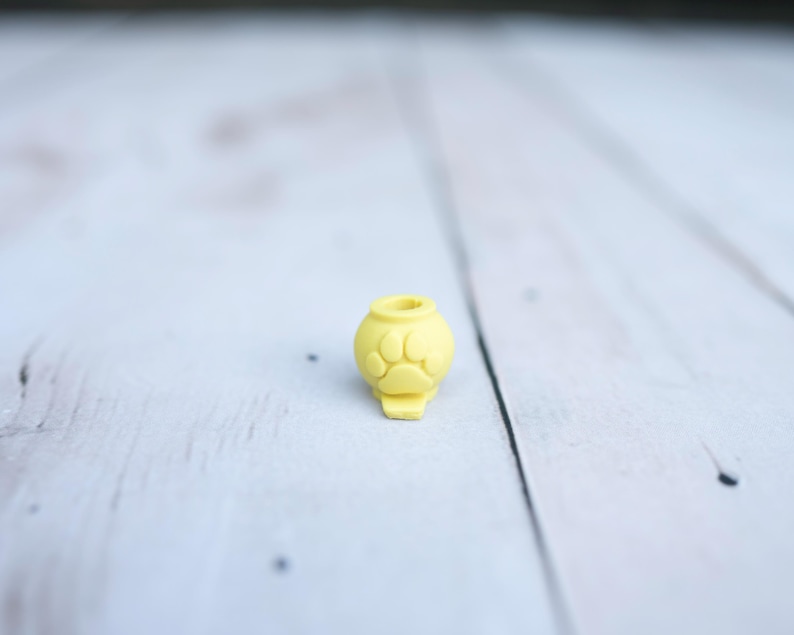 May include: A small, yellow, spherical bead featuring a raised paw print design on its front surface. The bead has a circular opening at the top and a small, flat, protruding base at the bottom, designed for threading onto a cord or string.