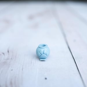 May include: A small, light blue, spherical bead featuring a raised paw print design on its front surface. The bead has a central hole at the top, designed for threading onto a cord or string, and a flat base for stability when placed on a surface.