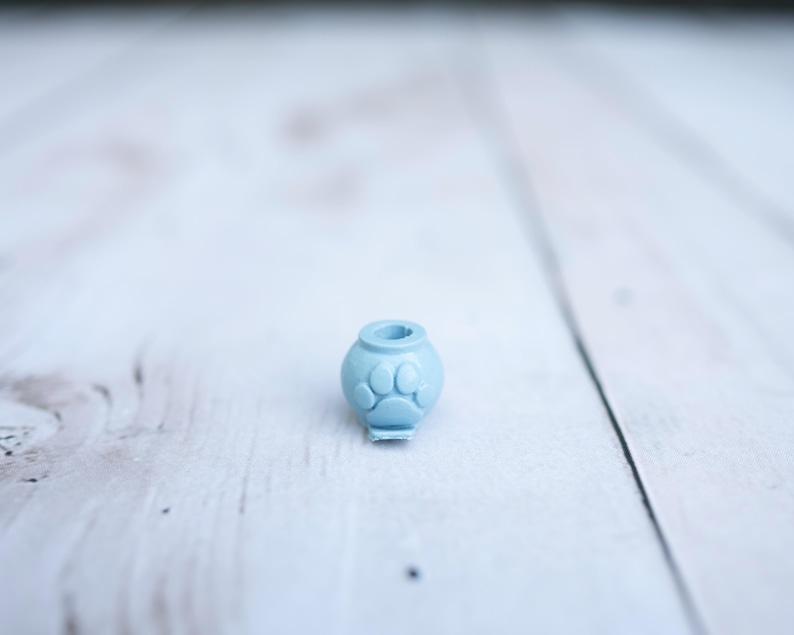 May include: A small, light blue, spherical bead featuring a raised paw print design on its front surface. The bead has a central hole at the top, designed for threading onto a cord or string, and a flat base for stability when placed on a surface.
