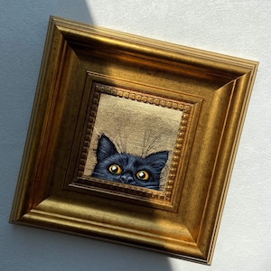 May include: A framed painting of a black cat peeking out with bright yellow eyes. The cat's head is centered within a gold-colored, ornate frame. The background of the painting is a textured gold color, and the frame is rectangular.