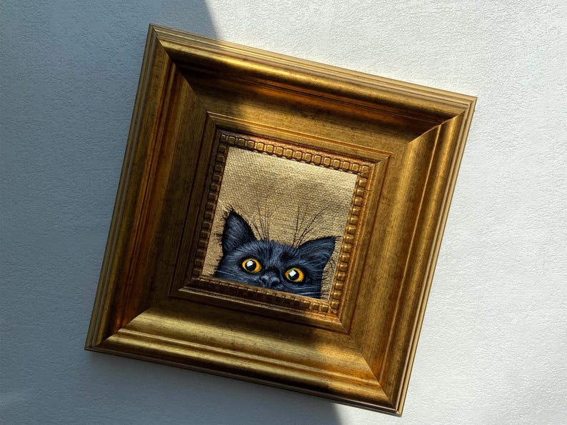 May include: A framed painting of a black cat peeking out with bright yellow eyes. The cat's head is centered within a gold-colored, ornate frame. The background of the painting is a textured gold color, and the frame is rectangular.