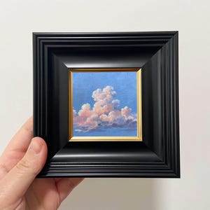 May include: A small framed painting depicting a cloudscape. The artwork shows a blue sky with fluffy, pink and white clouds. The painting is set within a gold-coloured frame and a black, ornate outer frame.