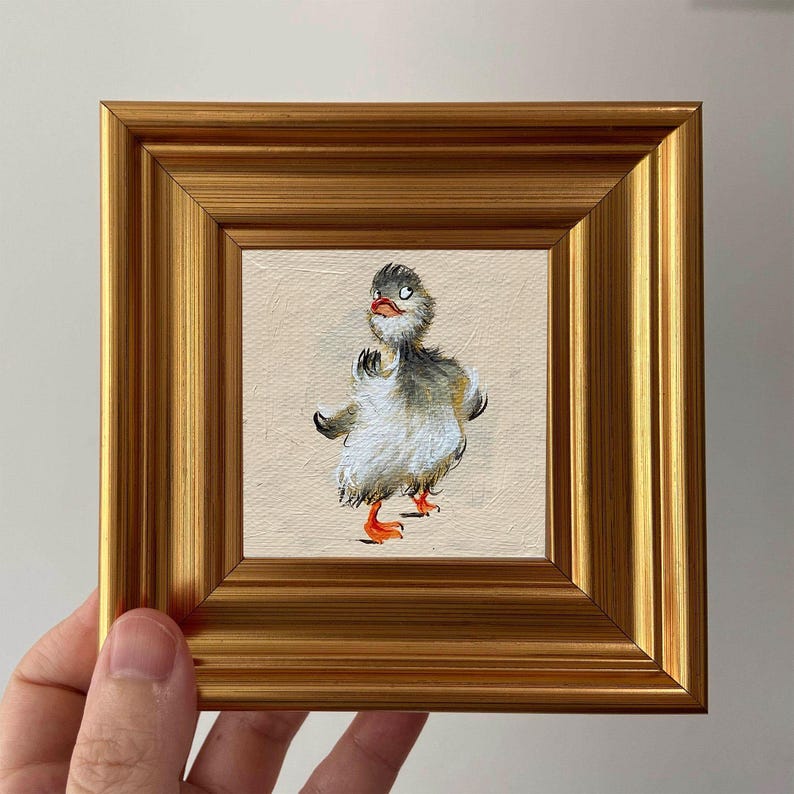 May include: A framed painting of a cartoon duckling with gray, white, and orange details. The duckling is centered on a beige background, framed by a gold-colored, square frame. The artwork is held by a hand.